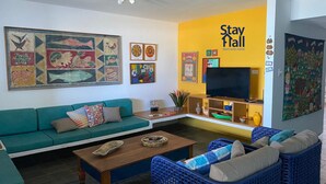 Un soggiorno con un divano verde, poltrone in vimini blu, un tavolino in legno e una TV su un mobile. Sono presenti decorazioni colorate alle pareti e un cartello con la scritta 'Stay Hall'.