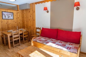 A wooden interior with a sofa, a dining table, and two chairs. There are two wall-mounted lamps.