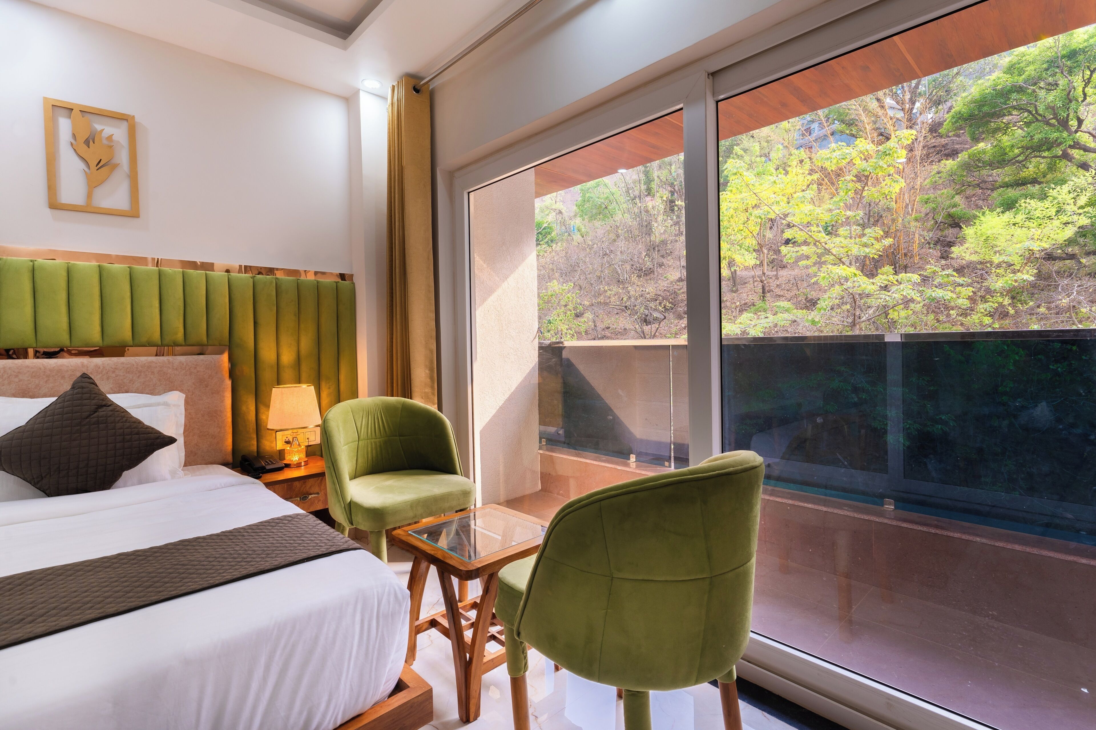 A hotel room with a bed, a green armchair, a small table, and a large window offering a view of trees.