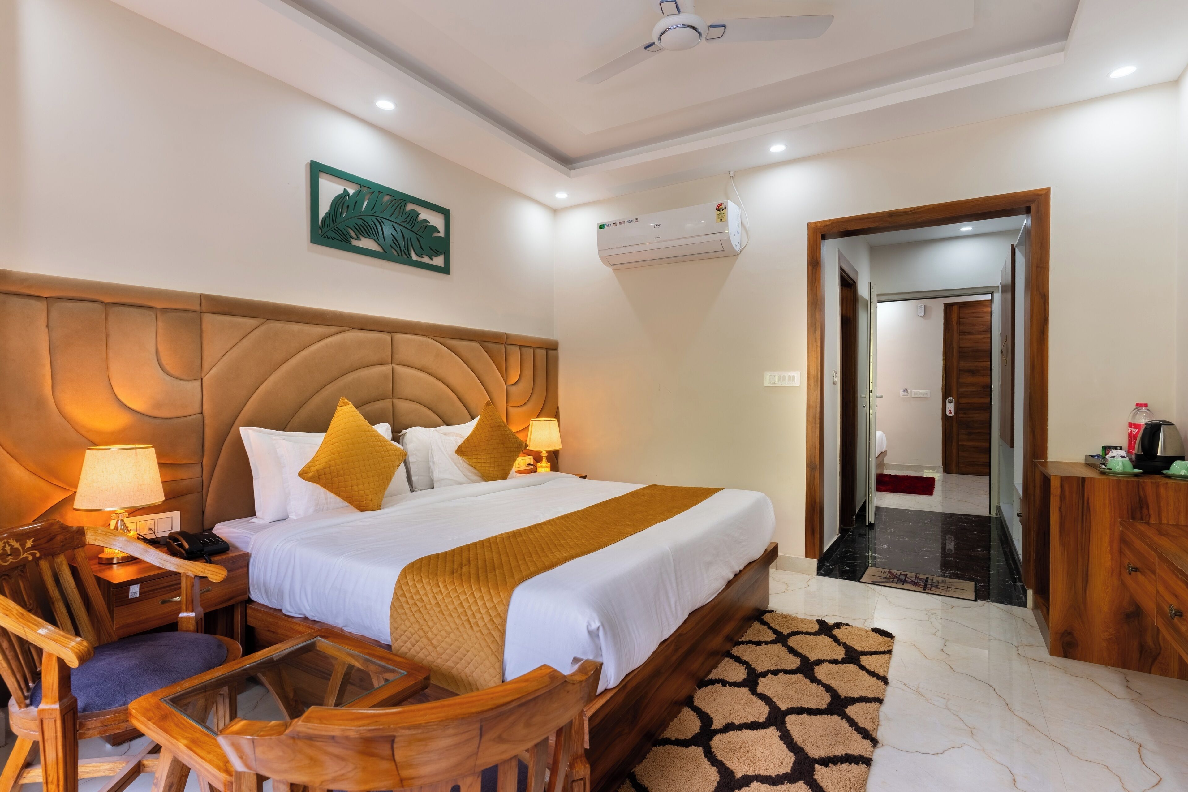A hotel room with a large bed, wooden headboard, two bedside tables with lamps, a wooden chair, a desk, a mirror, and a door leading to another room.