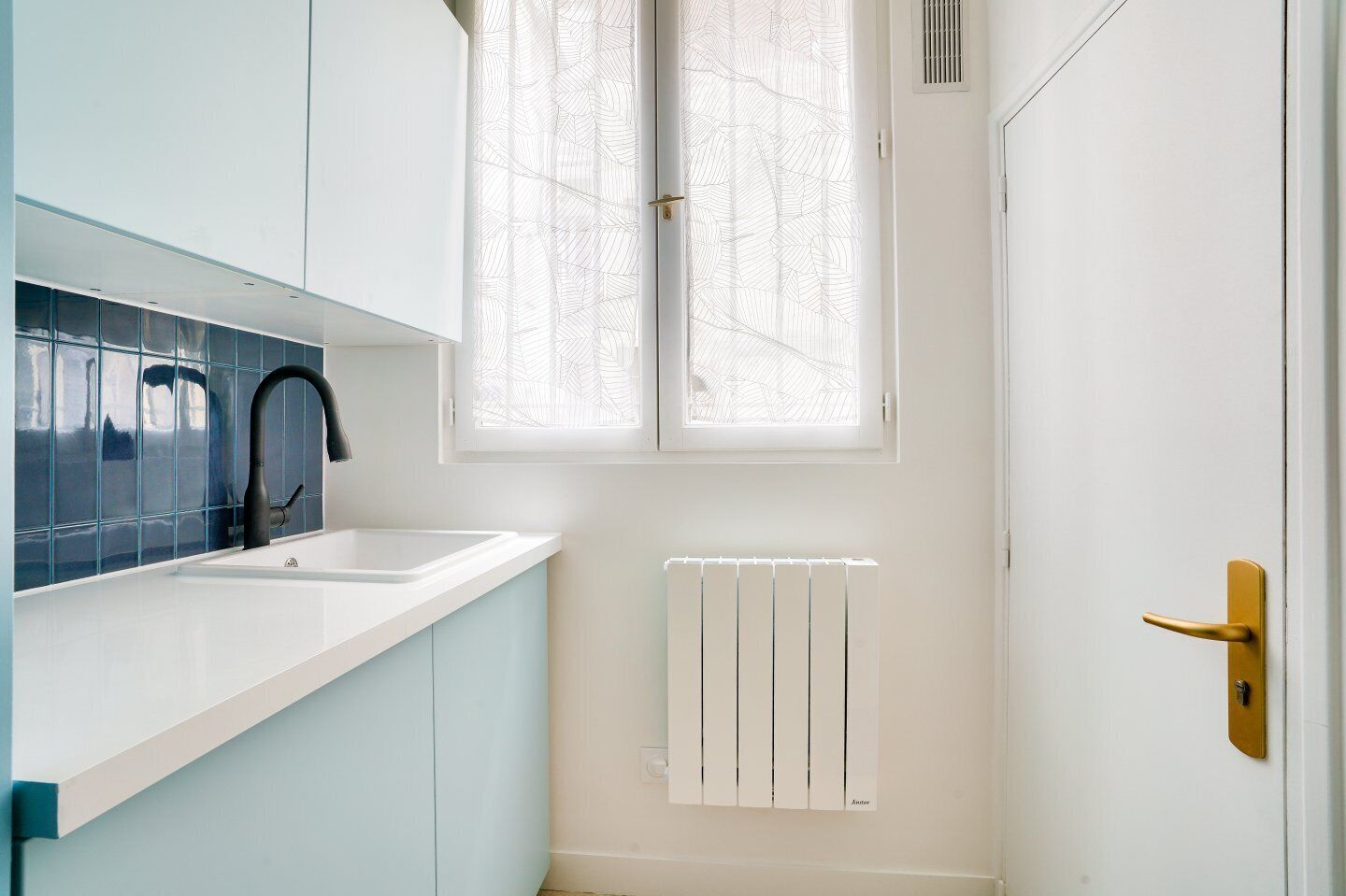 Una cocina con cubierta blanca, un grifo negro, una ventana con persianas de diseño de hojas y un radiador blanco.