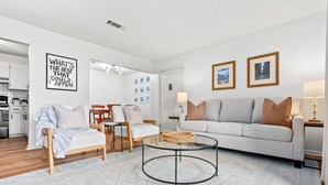 A modern living room with a grey sofa, a round coffee table, and a white armchair. There are framed pictures on the wall and a kitchen area in the background. - Gay Street Retreat - Walk to Campus & Downtown (Auburn)
