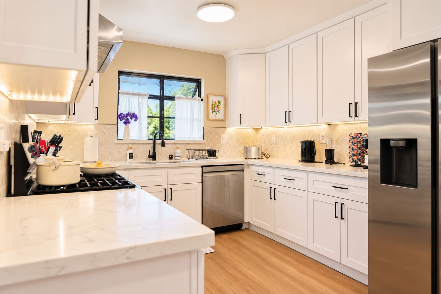 A modern kitchen with white cabinetry, a stainless steel refrigerator, a dishwasher, and a window with curtains.
