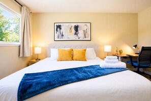 Ett hotellrum med en säng, två sänglampor, ett skrivbord med en stol, ett fönster med gardiner och en tavla på väggen.