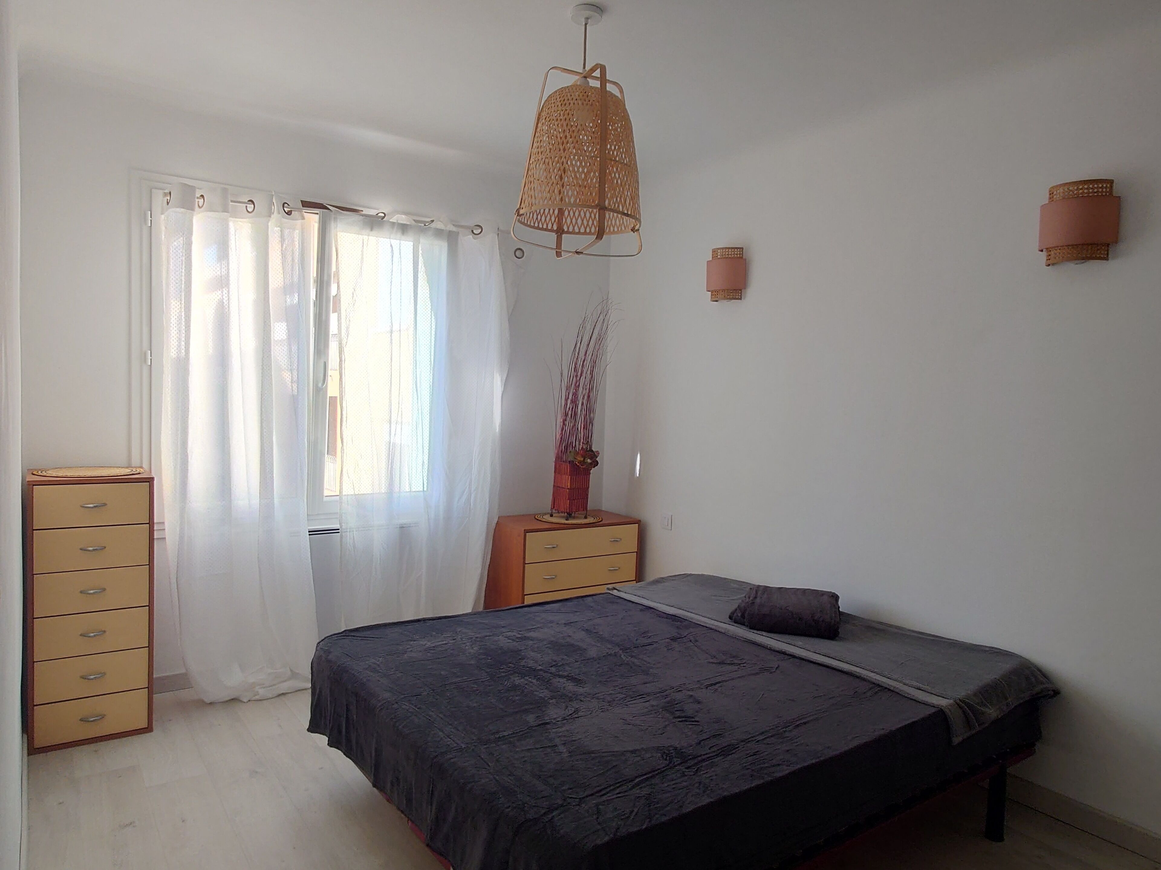 Een slaapkamer met een bed, een dressoir, een raam met dunne gordijnen, een hanglamp en een wandlamp.