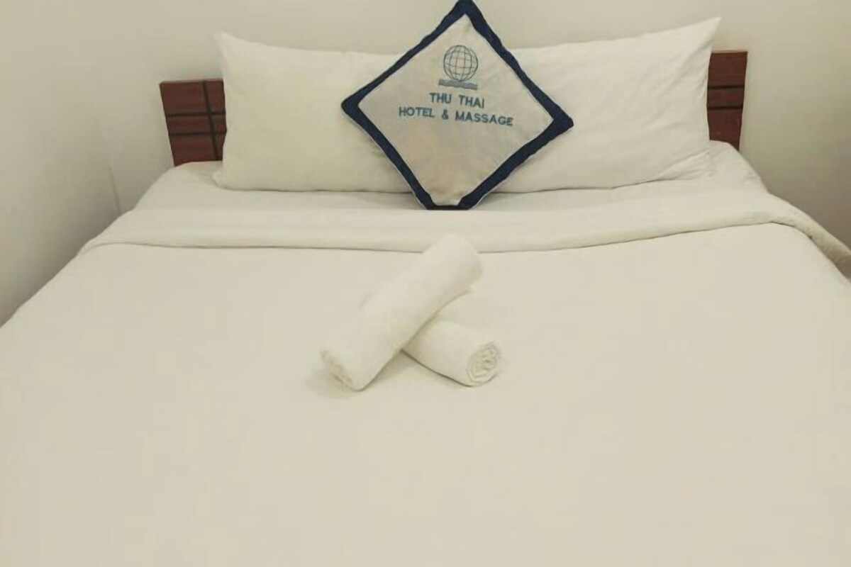 Een hotelbed met een kussen waarop 'THU THAI HOTEL & MASSAGE' staat en een netjes gevouwen witte handdoek.