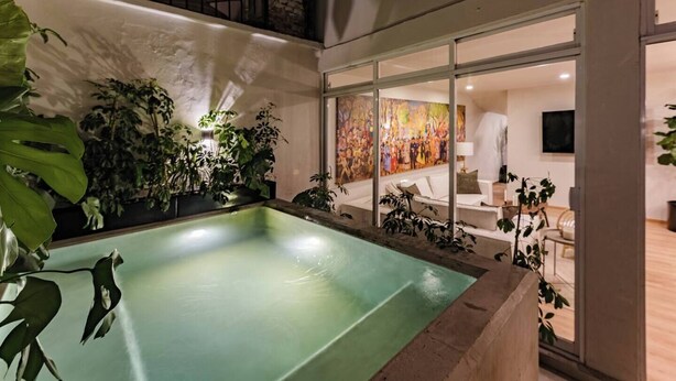 A modern indoor pool area with a glass enclosure, surrounded by lush greenery and a vibrant mural.