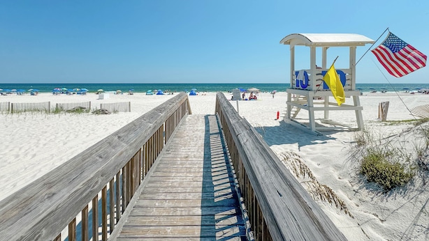 Ein hölzerner Steg führt zu einer Rettungswache mit einer US-Flagge, Strandmuscheln und Sand.
