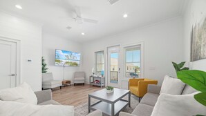 Ein modernes Wohnzimmer mit einem grauen Sofa, einem weißen Sessel, einem Couchtisch und einem gelben Sessel. Ein Fernseher ist an der Wand montiert, ein Deckenventilator ist vorhanden und ein großes Fenster mit Jalousien.