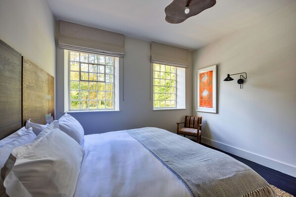 A neatly made bed with white linens, two windows with blinds, a chair, and a framed artwork on the wall.