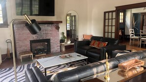 A modern living room with a fireplace, leather sofa, and a flat-screen TV mounted on the wall. - Historical home w gardens in Unique North Lawrence. ACROSS FROM TRAIN DEPOT (Lawrence)