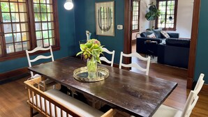 A dining area with a wooden table, white chairs, and a vase of flowers. There are large windows with wooden frames, and a dark blue wall. - Historical home w gardens in Unique North Lawrence. ACROSS FROM TRAIN DEPOT (Lawrence)