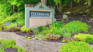 Segnaletica per 'CLEARBROOK' circondata da un giardino curato con diverse piante e un muro di sostegno in pietra.