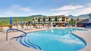 Piscina di un resort con lettini e ombrelloni, circondata da un edificio con numerosi balconi e una montagna sullo sfondo.