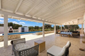 Un patio exterior con mobiliario de mimbre, una mesa de piedra y vistas a una piscina y zonas verdes.