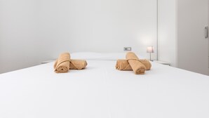 Habitación de hotel con una cama grande, dos toallas enrolladas, una mesita de noche y una lámpara fijada a la pared.