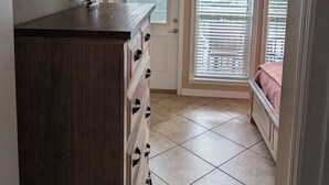 A bedroom with a wooden dresser, a flat-screen TV mounted on the wall, and a window with blinds. - Beach Beau (Long Beach)