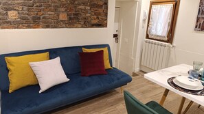 A room with a blue sofa, a white and yellow pillow, a red pillow, a wooden table with a white plate, and a window with a white curtain.