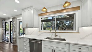 Cocina moderna con armarios blancos, lavavajillas de acero inoxidable, un amplio ventanal con vistas e iluminación colgante.
