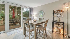 A dining area with a wooden table and chairs, a glass-top table, a lamp, and a wall-mounted shelf with decorative items.