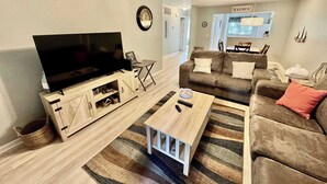 A living room with a flat-screen TV on a wooden stand, a beige sofa, a coffee table, and a striped rug. - Pet Friendly Beach Villa 2 Bed 2 Bath #7105 TreeTops (Hilton Head Island)