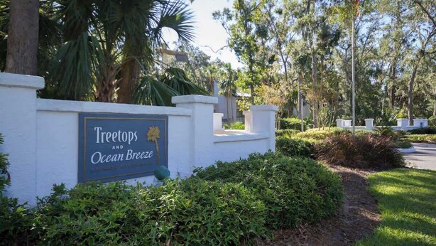A sign for 'Treetops and Ocean Breeze' with a white wall and greenery in the background. - Pet Friendly Beach Villa 2 Bed 2 Bath #7105 TreeTops (Hilton Head Island)