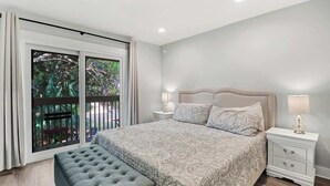 Un dormitorio con una cama grande, un banco, una mesita de noche y una ventana con vista a áreas verdes.