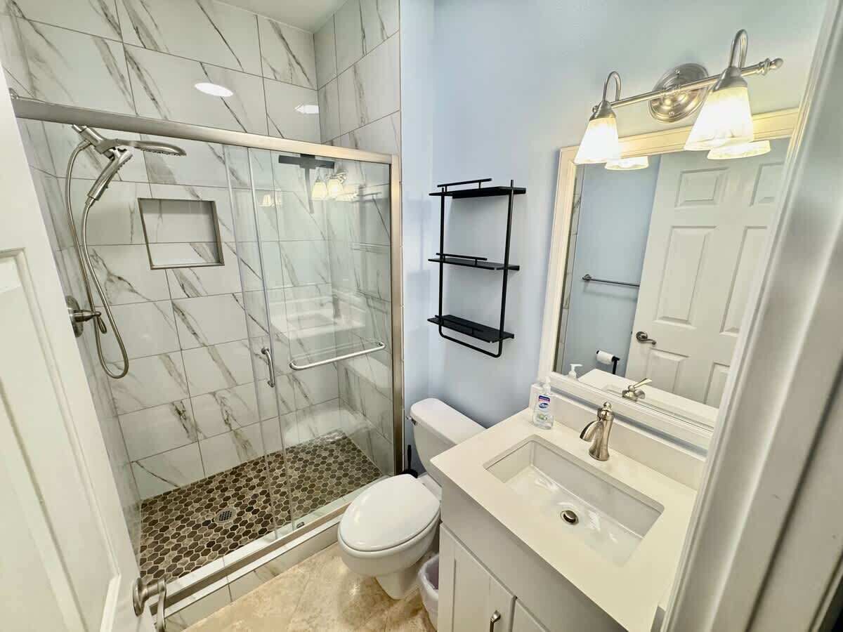 Une salle de bain avec une douche à parois de verre, des toilettes blanches, un lavabo avec un miroir et une étagère noire.