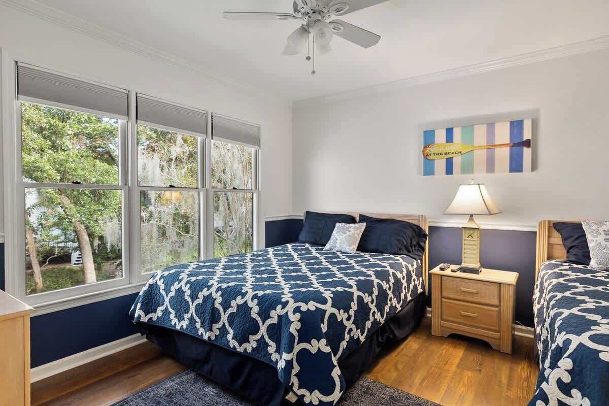 A bedroom with a large window, a ceiling fan, a bed with a patterned bedspread, a bedside table with a lamp, and a wall art piece.