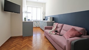 Un salon moderne avec un canapé, un téléviseur à écran plat fixé au mur et un coin cuisine à l’arrière-plan.