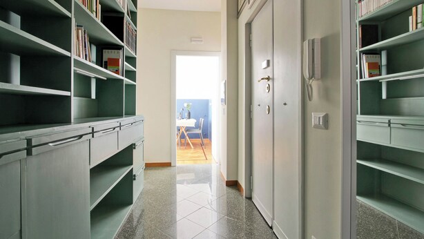 Un couloir avec des étagères vertes remplies de livres, une porte fermée avec un téléphone, et une vue sur un coin repas avec une table et des chaises.