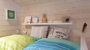 Une chambre avec des murs en bois, un lit recouvert d’une literie aux couleurs vives, une étagère remplie de livres et une statue de Bouddha, ainsi qu’une plume posée sur le lit.