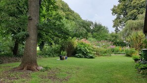 Un jardin luxuriant, avec une grande variété de plantes et d’arbres, une pelouse bien entretenue et un grand tronc d’arbre au premier plan.