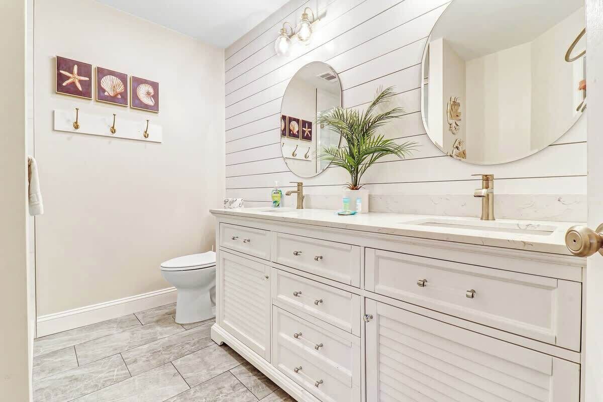 A bathroom with a white vanity, a round mirror, and a toilet. There are decorative wall art pieces and a plant on the vanity.