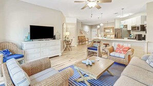 Una sala de estar moderna con un televisor de pantalla plana, muebles de mimbre, un ventilador de techo y una cocina con microondas y refrigerador.