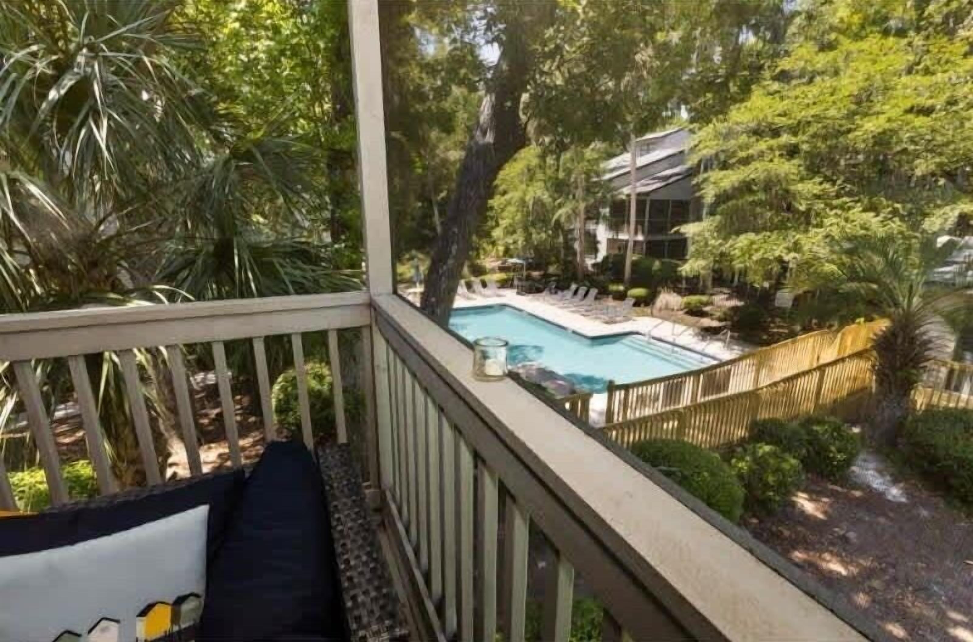 Un área de piscina con terraza de madera, sillas de descanso y vista a una casa al fondo.