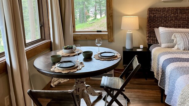 A cozy room with a round dining table set for two, a bed with a striped duvet, a bedside lamp, and a view of the outdoors through a window. - Ski-In/Ski-Out • Romantic Studio • Near Trails (Angel Fire)