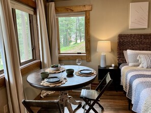 A cozy room with a round dining table set for two, a bed with a striped duvet, a bedside lamp, and a view of the outdoors through a window.