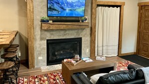 A living room with a fireplace, a mounted TV, a wooden coffee table, and a sofa. - Ski-In/Ski-Out • Romantic Studio • Near Trails (Angel Fire)