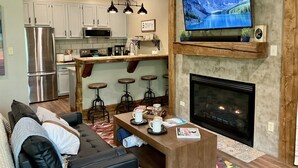 A modern living room with a fireplace, a large TV mounted above it, and a kitchen area with white cabinets and a stainless steel refrigerator. - Ski-In/Ski-Out • Romantic Studio • Near Trails (Angel Fire)