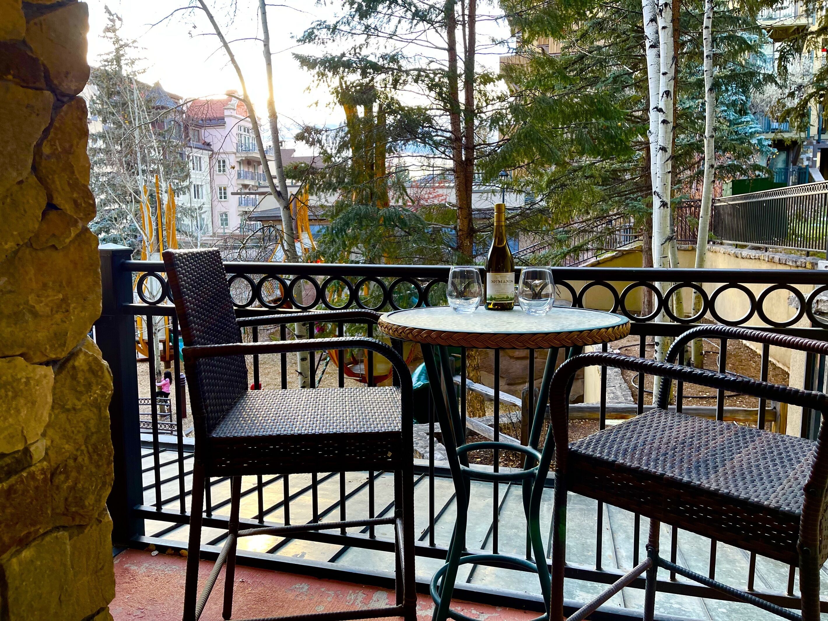 En balkon med fletstole og et bord, med udsigt til en legeplads og bygninger.