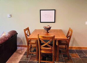 Een eetgedeelte met een houten tafel en stoelen, een kunstwerk in een lijst aan de muur en een mand met snacks op de tafel.