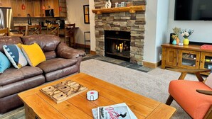 Un salon chaleureux avec une cheminée en pierre, un canapé en cuir, une table basse en bois et un téléviseur à écran plat fixé au mur.