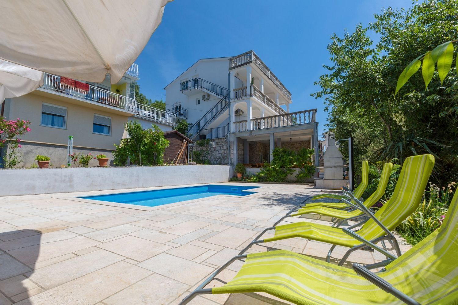 A pool area with a rectangular pool, a covered seating area with yellow striped chairs, and a building with multiple balconies and a stone facade.