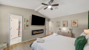 A bedroom with a large bed, a ceiling fan, a flat-screen TV, a fireplace, and artwork on the walls.