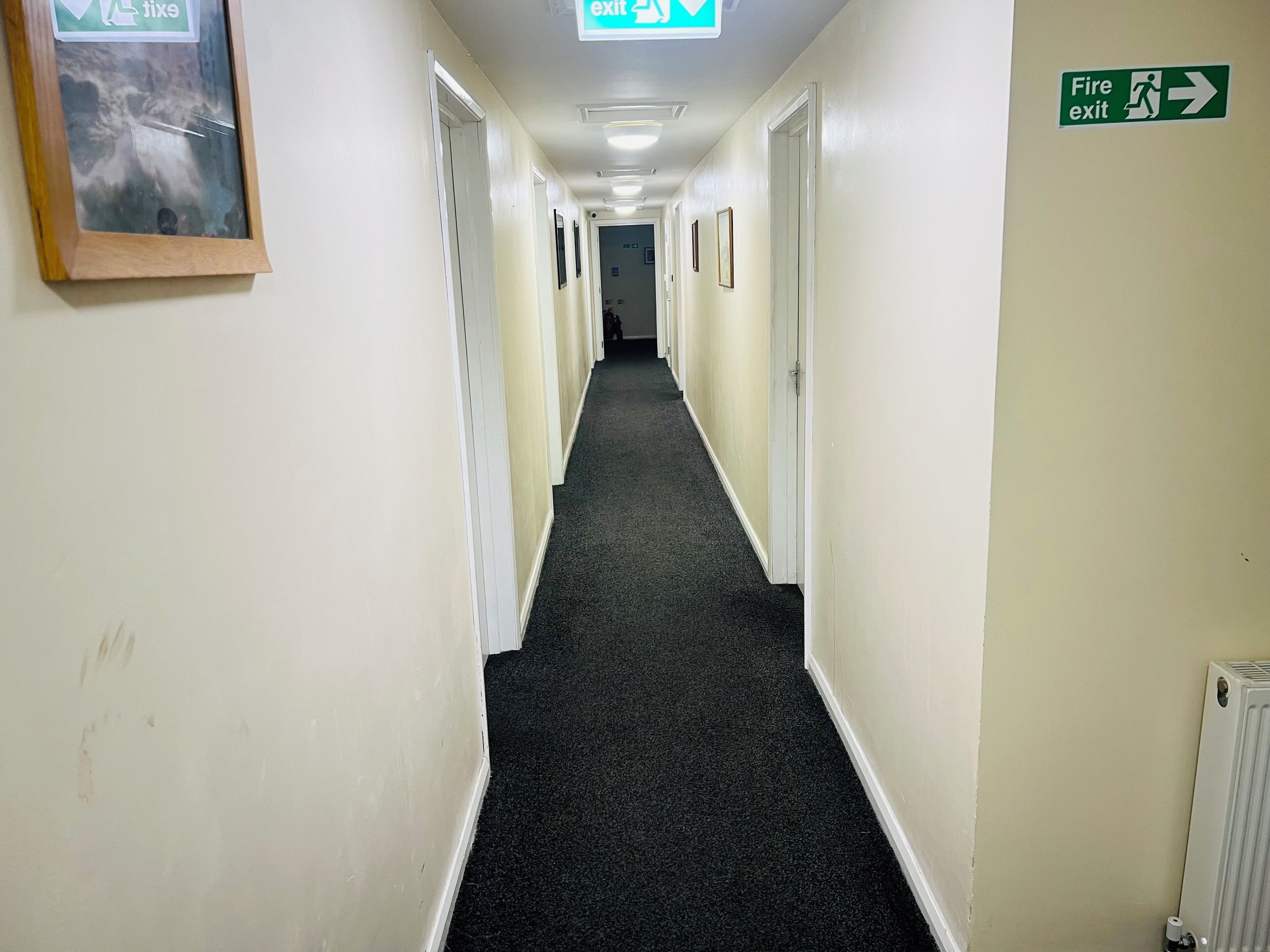 A hallway with carpeted floors, white walls, and doors on both sides. There are exit signs and a fire exit sign on the walls.