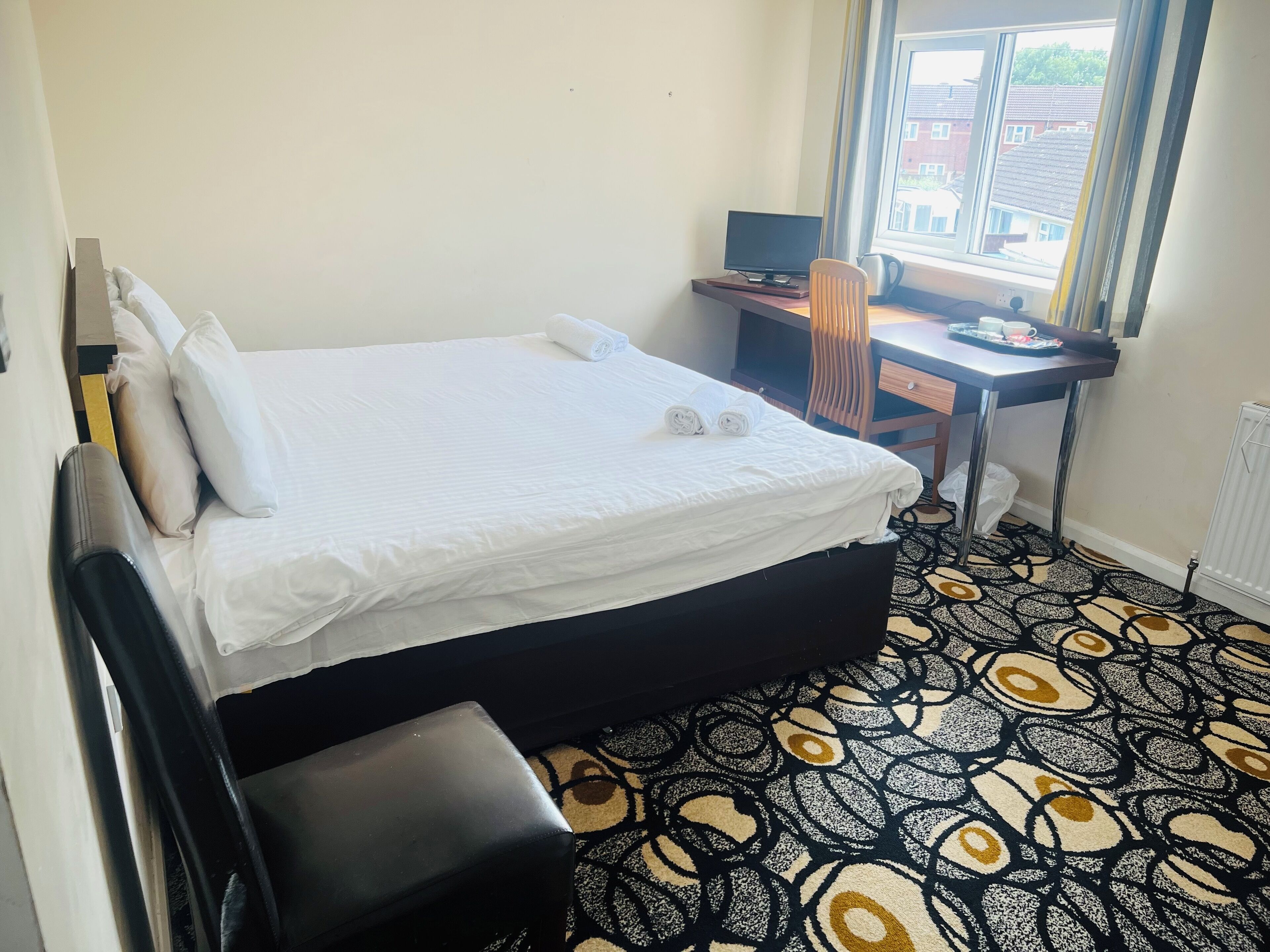 A neatly made bed with white linens, a small desk with a computer, and a patterned carpet.