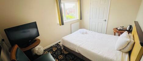 A hotel room with a bed, a television, a chair, a small table with a kettle, and a window with curtains.