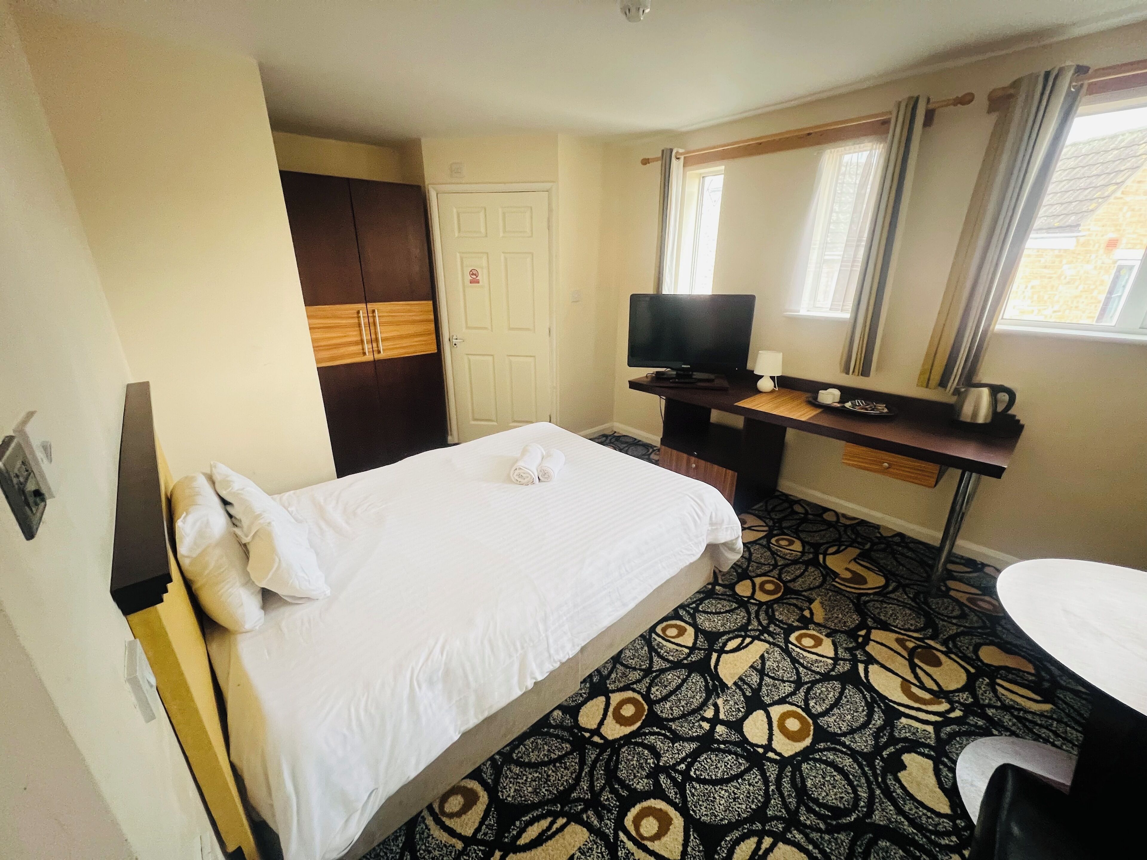 A hotel room with a bed, a desk with a television, a small table with a cup and a kettle, and a patterned carpet.
