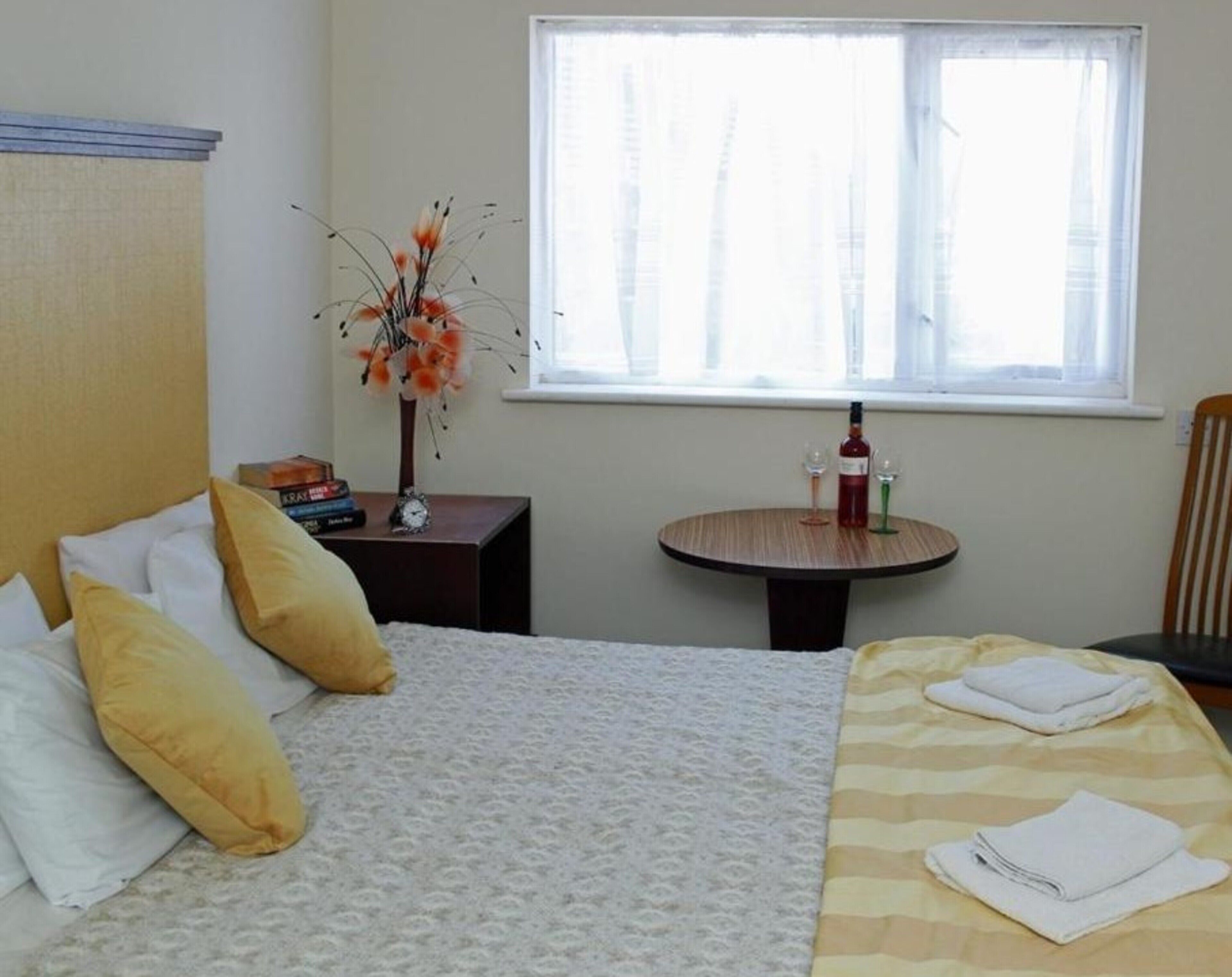 A hotel room with a bed, a small table with a bottle and glasses, a vase with flowers, and a window with blinds.